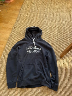 Mörkblå hoodie Abercrombie & Fitch - Snygg mörkblå hoodie från Abercrombie & Fitch med vit logga och text på bröstet. Klassisk känguruficka framtill och justerbar huva med vita snören. Perfekt för en avslappnad och trendig stil.