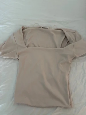 Beige topp med fyrkantig ringning - Säljer en stilren beige topp med korta ärmar och fyrkantig ringning. Toppen har en enkel och clean design som passar perfekt till jeans eller kjol. Materialet känns mjukt och stretchigt, vilket gör den bekväm att bära hela dagen.