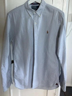 Randig skjorta från Polo Ralph Lauren - Klassisk långärmad skjorta från Polo Ralph Lauren i vitt och ljusblått randigt bomullstyg. Skjortan har button-down krage, broderad logga på bröstet och knappar framtill. Perfekt för en clean och stilren look.