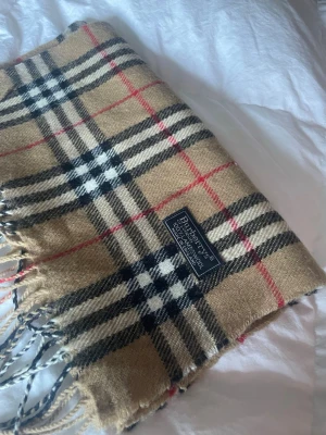 Klassisk beige rutig halsduk Burberry - Säljer en ikonisk beige halsduk från Burberry med klassiskt rutmönster i svart, vitt och rött. Halsduken är tillverkad i 100% lammull och har fransar i ändarna. Perfekt accessoar för att lyfta din outfit med en tidlös touch.