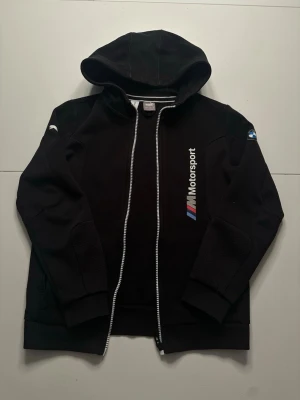 Svart Puma BMW M Motorsport hoodie - Säljer en svart hoodie med dragkedja från BMW M Motorsport. Hoodien har tryckta loggor på bröstet och ärmen, samt en huva och ribbade muddar. Materialet känns mjukt och sportigt, perfekt för dig som gillar motorsport och BMW. Den är i super bra skick och har använts enstaka gånger. Det står att den passar från 11-12 år men den är liten i storkleken så snarare 6-10 år.👍Bara höra av dig om du har några funderingar!