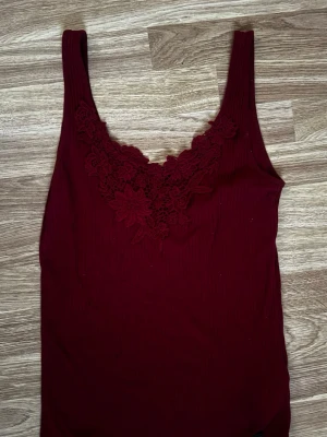 Vinröd linne med spetsdetaljer från Hollister  - Snygg vinröd linnetopp från Hollister med breda axelband och v-ringning dekorerad med blommig spets. Perfekt att styla till jeans eller kjol för en trendig look. Materialet känns mjukt och följsamt mot huden. 