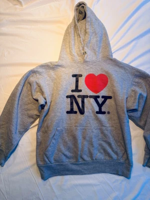 Grå hoodie med I ❤️ NY-tryck - Säljer en grå hoodie med det klassiska I ❤️ NY-trycket på bröstet. Hoodien har stor luva, magficka och ribbade muddar. Perfekt för dig som gillar streetwear och vill ha en ikonisk look. Materialet känns mjukt och bekvämt. storlek S 