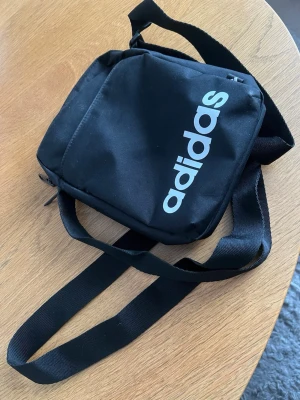 Svart axelväska från Adidas - Snygg svart axelväska från Adidas med justerbar rem och stort vitt logotryck på framsidan. Väskan har en fyrkantig form, dragkedja upptill och är tillverkad i slitstarkt tygmaterial. Perfekt för att bära det viktigaste när du är på språng.