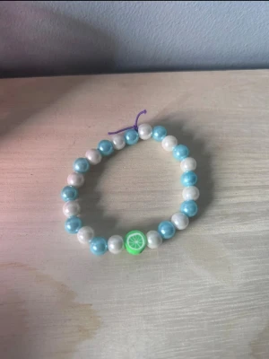 Pärlarmband med limepärla - Säljer ett färgglatt armband med vita och turkosa pärlor samt en grön limeformad pärla i mitten. Trädd på elastisk lila tråd, perfekt för dig som gillar unika accessoarer med fruktig touch.