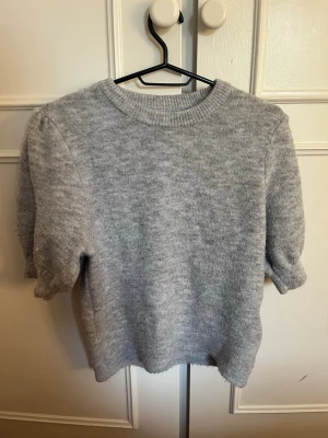 Grå stickad tröja från Gina Tricot - Mysig grå stickad tröja från Gina Tricot med korta ärmar och rund halsringning. Tröjan har en enkel och stilren design, perfekt att matcha med jeans eller kjol. Materialet känns mjukt och bekvämt mot huden.