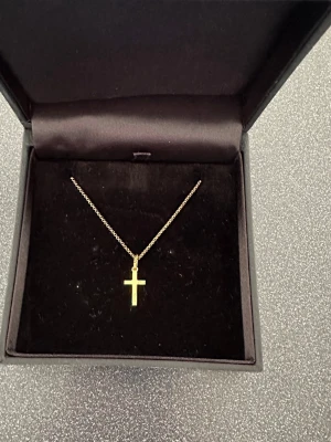 Guldigt halsband med korshänge - Guldpläterat halsband i guldton med ett stilrent korshänge. Kedjan är tunn och elegant, perfekt för dig som gillar minimalistiska smycken. Levereras i en svart ask.