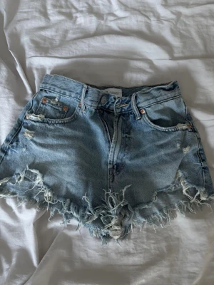Blå jeansshorts med fransig kant - Säljer ett par blå jeansshorts med fransig och sliten kant. Shortsen har klassisk femficksdesign, knapp och dragkedja framtill samt slitna detaljer för en cool look.