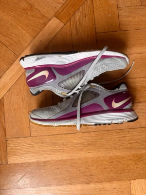 Nike grå och lila sneakers - Snygga sneakers från Nike i grått och lila med vit sula och klassisk swoosh-logga på sidan. Skorna har meshpaneler för bra ventilation och detaljer i plast för extra stöd. Perfekta för dig som gillar sportig stil och vill ha bekväma skor till vardagen.