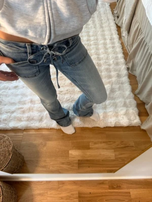 Blå snörade jeans från Gina Tricot - Säljer ett par ljusblå jeans från Gina Tricot med snörning i midjan och utsvängda ben. Jeansen har coola detaljer med metallöljetter och är i klassisk denim. Perfekta för dig som gillar en unik twist på dina jeans!