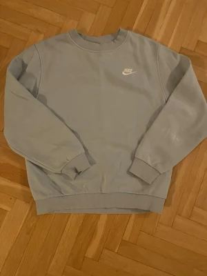Ljusblå sweatshirt från Nike - Ljusblå sweatshirt från Nike med klassisk rund halsringning och diskret Nike-logga broderad i vitt på bröstet. Tröjan har ribbade muddar vid ärmslut och nederkant. Perfekt för en avslappnad och sportig stil. Har en liten skavank på ena armen