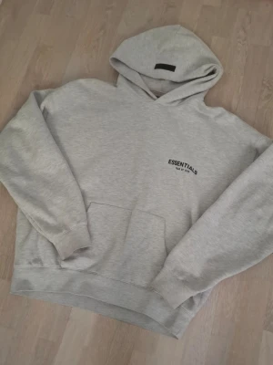 Grå Essentials Fear of God hoodie - Snygg ljusgrå hoodie från Essentials Fear of God med svart logga på bröstet och stor text på ryggen. Klassisk känguruficka framtill och huva. Tillverkad i mjukt sweatshirtmaterial, perfekt för en avslappnad streetwear-look.