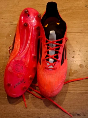 Röda Adidas X Speedflow fotbollsskor - Säljer ett par röda Adidas X Speedflow fotbollsskor med svarta detaljer och snörning. Skorna har dobbar för spel på gräs och är tillverkade i ett lätt syntetmaterial som ger bra grepp och känsla på planen. Perfekta för dig som vill ha snabbhet och kontroll.
