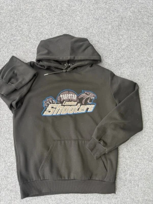 Svart hoodie Trapstar London Shooters - Svart hoodie med stor broderad logga på bröstet där det står 'Trapstar London Shooters' och motiv av en panter. Hoodien har huva med snörning och en klassisk magficka. Perfekt för dig som gillar streetwear och vill sticka ut.