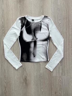 Vit långärmad topp med tryck - Trendig vit långärmad topp med svart grafiskt tryck framtill som ger en edgy look. Figurnära passform och rund halsringning. Perfekt för dig som vill sticka ut med din stil.