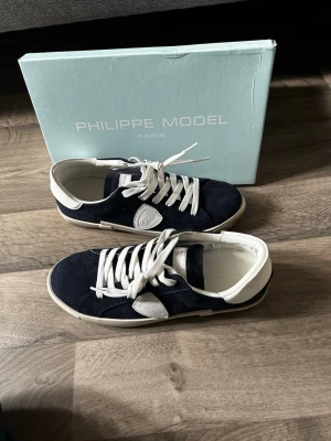 Philippe Model blåa sneakers - Mörkblå sneakers från Philippe Model med vita detaljer och vita skosnören. Skorna har en låg silhuett, vit sula och är tillverkade i mocka och läder. Klassisk Philippe Model-logga på sidan och bekväm innersula. Perfekta för en stilren och avslappnad look.