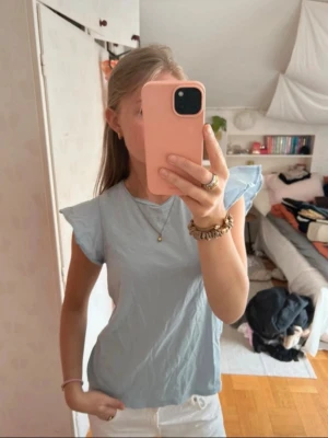 Ljusblå topp med volangärmar - Snygg ljusblå topp med korta volangärmar och rund halsringning. Perfekt för en trendig och avslappnad look. Tunt och lätt material som känns skönt mot huden.