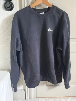 Svart sweatshirt från Nike - Klassisk svart sweatshirt från Nike med vit broderad logga på bröstet. Tröjan har rund halsringning, långa ärmar och ribbade muddar vid ärmslut och nederkant. Perfekt för en avslappnad och sportig stil.