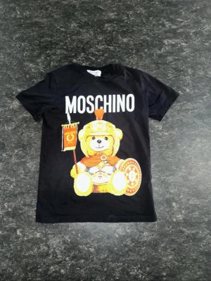 Svart Moschino t-shirt med björntryck - Cool svart t-shirt från Moschino med stort tryck av en björn utklädd till romersk soldat på framsidan. T-shirten har rund hals och korta ärmar. Perfekt för dig som vill sticka ut med en lekfull och trendig look.