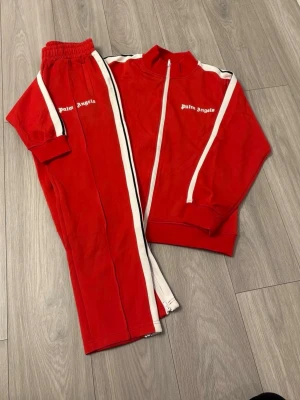 Rött loungewear-set från Palm Angels - Palm Angels rött loungewear-set med vit dragkedja och vita ränder längs ärmar och ben. Setet består av en zip-jacka och matchande byxor, båda med logga på bröst och lår. Tillverkat i mjukt material, perfekt för chill dagar hemma.