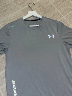 Grå tränings-tshirt från Under Armour - Snygg grå tränings-tshirt från Under Armour med rund hals och korta ärmar. Logga på bröstet och text på sidan. Tillverkad i ett lätt och snabbtorkande material som passar perfekt till gymmet eller löprundan.