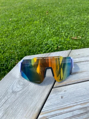 Multifärg sportglasögon - Säljer ett par sportiga solglasögon med stora spegelglas i guld, blå och gröna toner. Bågarna är vita och modellen har en futuristisk, kantig form som täcker mycket av ansiktet. Perfekta för dig som vill sticka ut i sommar och skydda ögonen mot solen.