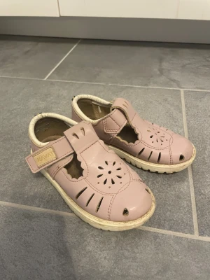 Rosa sandaler med kardborre - Söta rosa sandaler i läder med blommigt utskuret mönster på ovandelen och vit sula. Kardborreband gör dem enkla att ta på och av. Perfekta för vår och sommar, med en bekväm passform och klassisk design.