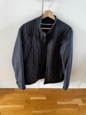 Svart quiltad jacka med ståkrage medium - Snygg svart quiltad jacka med ståkrage och dragkedja framtill. Jackan har ett rutmönster över hela ytan och är tillverkad i ett lätt, glansigt material. Perfekt för dig som vill ha en stilren och enkel jacka till vardags.