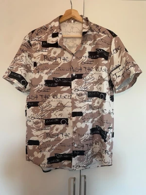 Beige kortärmad skjorta med textmönster - Säljer en beige och brun kortärmad skjorta med svart och vit graffiti-inspirerat textmönster. Skjortan har klassisk krage och knäppning framtill. Perfekt för dig som gillar streetwear och unika prints.