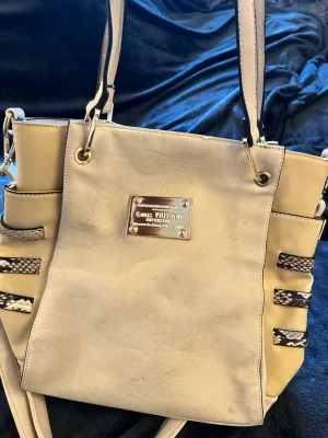 Beige handväska med ormmönstrade detaljer - Snygg beige handväska i konstläder med guldiga metalldetaljer och ormmönstrade band på sidorna. Väskan har både handtag och axelrem samt ett rymligt innerfack med tryckt tygfoder. Perfekt för dig som gillar trendiga accessoarer.
