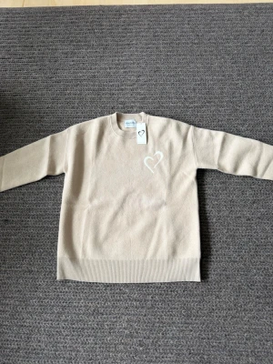 Beige stickad tröja med hjärta - Beige stickad tröja med rund halsringning och ribbade muddar. På bröstet finns ett vitt broderat hjärta som detalj. Tröjan har en enkel och stilren design, perfekt för en avslappnad look.