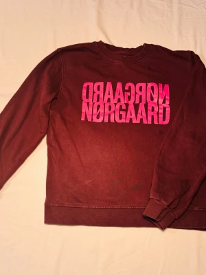 Vinröd sweatshirt från Mads Nørgaard - Vinröd sweatshirt med rund hals och långa ärmar från Mads Nørgaard. Framsidan har en stor rosa logotyp i tryck. Tröjan har ribbade muddar vid ärmslut och nederkant. Tillverkad i mjukt bomullsmaterial för en skön känsla. Storlek 14 - XS. Har en liten fläck därav priset 
