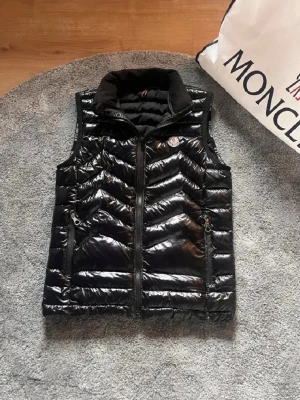 Svart dunväst från Moncler - Snygg svart dunväst från Moncler med glansig finish och hög krage i mjukt material. Västen har dragkedja framtill, två sidofickor med dragkedja och Moncler-logga på bröstet. Perfekt för lager på lager och streetstyle.