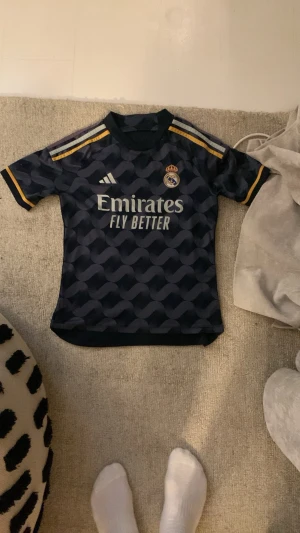 Real Madrid bortatröja Vini Jr #7 - Snygg mörkblå Real Madrid bortatröja med gult tryck och Vini Jr nummer 7 på ryggen. Tröjan har korta ärmar, gulddetaljer på axlar och ärmslut samt klubbmärke och sponsorlogga på bröstet. Tillverkad i lätt och ventilerande funktionsmaterial.