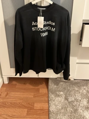 Svart sweatshirt Acne Studios - Svart sweatshirt från Acne Studios med vit text 'Acne Studios STOCKHOLM 1996' på bröstet. Klassisk rund halsringning och avslappnad passform. Perfekt för en stilren och trendig look.
