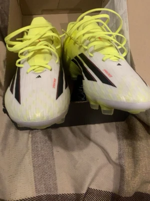 Adidas F50 vita och gula fotbollsskor - Säljer ett par Adidas F50 fotbollsskor i vitt och neongult med svarta detaljer och röda F50-tryck på sidan. Skorna har klassisk snörning och dobbar för spel på gräs. Lätta och smidiga i syntetmaterial, perfekta för dig som vill ha speed på planen.