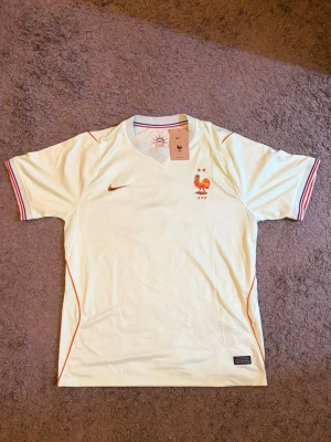 Nike fotbollströja Frankrike -  ljusgul fotbollströja från Nike med Frankrikes landslagsemblem och två stjärnor på bröstet. Tröjan har röda detaljer, Dri-Fit-material. Bra för träning eller match.