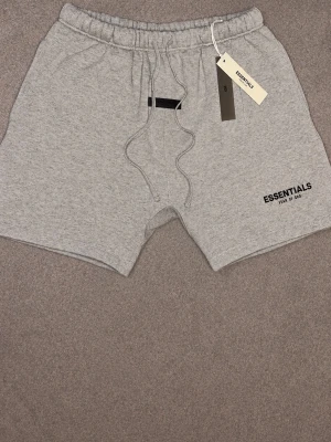 Grå shorts från Essentials Fear of God - Snygga grå shorts från Essentials Fear of God med snörning i midjan och logga tryckt på vänster ben. Tillverkade i mjuk bomull, perfekta för chill dagar eller streetwear-stil. Elastisk midja för skön passform.
