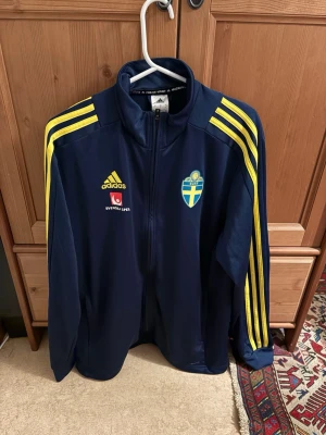 Blå svensk landslagsträningströja Adidas - Mörkblå träningströja från Adidas med Sveriges landslagsemblem och Svenska Spel-logga på bröstet. Tröjan har dragkedja, ståkrage och gula ränder längs ärmarna. Perfekt för fotbollsträning eller som supporterplagg.