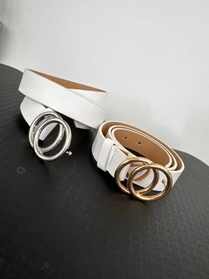Vita bälten med dubbelring-spänne - Två stilrena vita bälten med trendigt dubbelring-spänne, ett i silver och ett i guld. Bältena har en slät yta och passar perfekt till jeans eller kjol för en modern look. Materialet ser ut att vara syntet med en klassisk design.