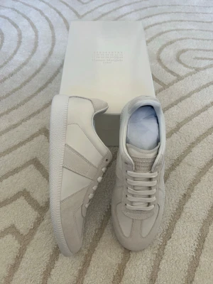 Vita sneakers från Maison Margiela - Stilrena vita sneakers från Maison Margiela med klassisk låg silhuett. Skorna har ovandel i läder och mocka, vita snören och en vit gummisula med mönstrad undersida. Perfekta för dig som gillar minimalistisk och clean stil.