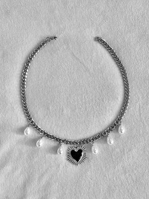 Silverhalsband med pärlor och hjärta - Snyggt halsband i silverfärgad u med droppformade vita pärlor och ett svart hjärthänge med solstråleliknande detaljer runt. Kedjan är chunky och ger ett trendigt intryck, perfekt för att lyfta din outfit med en cool och unik touch.