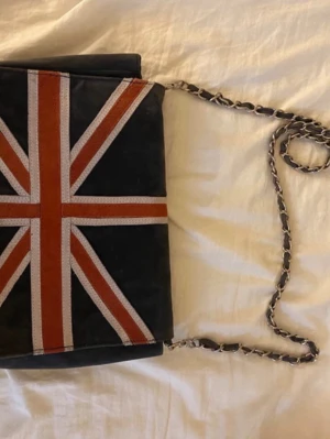 Svart axelväska från Gina Tricot - Stilren svart axelväska från Gina Tricot med kedjerem och lock. Väskan har en dekorativ rand i rött och grått på locket och är gjord i ett mjukt mockaliknande material. Perfekt för dig som vill ha en enkel men snygg accessoar.