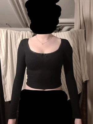 Svart croppad långärmad topp - Snygg svart croppad topp med långa ärmar och ribbad struktur. Toppen har en liten rosett och spetsdetaljer vid urringningen som ger en söt touch. Perfekt att matcha med höga jeans eller kjol.