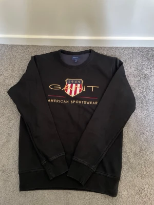 Svart sweatshirt från GANT - Säljer en svart sweatshirt från GANT med rund halsringning och tryck framtill. Tröjan har ett stort GANT-logotyptryck med amerikansk sköld och texten 'American Sportswear' i guld och rött. Klassisk och stilren modell i mjukt material.