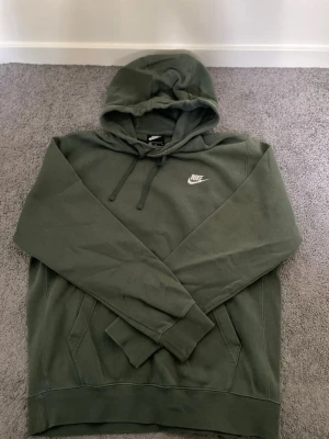 Olivgrön hoodie från Nike - Snygg olivgrön hoodie från Nike med klassisk logga broderad i vitt på bröstet. Hoodien har huva med dragsko, känguruficka framtill och ribbade muddar vid ärmar och nederkant. Tillverkad i mjuk bomullsblandning för skön känsla.