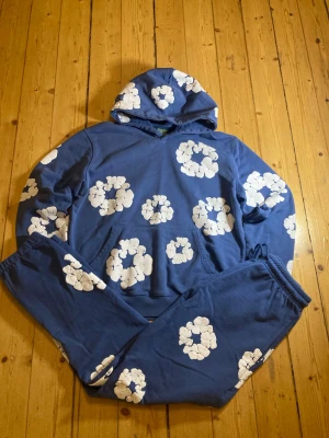 Blå denim tears tracksuit med vita blommor - Säljer en blå hoodie med huva och stora vita blommönster över hela tröjan. Hoodien har en avslappnad passform och är gjord i mjukt bomullstyg. Perfekt för dig som gillar färgglada och unika plagg med skön streetwear-vibe.