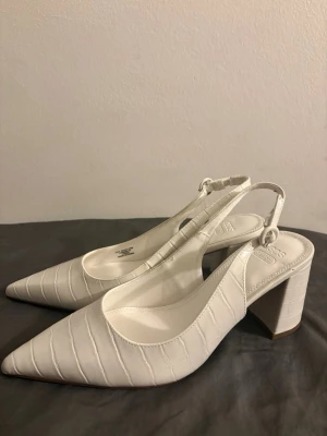 Vita klackar  - Säljer ett par vita pumps med spetsig tå och bred klack. Skorna har ett snyggt krokopräglat mönster och justerbar rem med rund spänne i hälen. Tillverkade i konstläder för en trendig look.