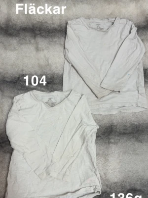 Två vita långärmade t-shirts - Två vita långärmade t-shirts. Klassisk rund halsringning och enkel design utan tryck eller detaljer. Perfekta som basplagg till vardags eller under tröjor.🌸 Fläckar. Storlek 104 