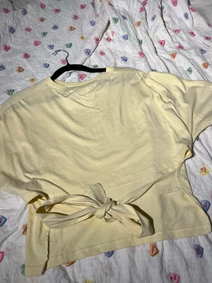 Topp med öppen rygg och knytning - Superfin ljusgul topp i bomull med korta ärmar och rund hals. Toppen har en snygg knyt-detalj i midjan i ryggen som ger en trendig look. Perfekt för dig som gillar en enkel men ändå unik stil.